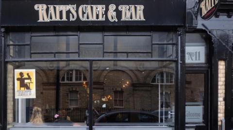 The exterior of Rapp's Cafe & Bar. It has a large glass window while the frames and walls are painted black with a logo in gold above.