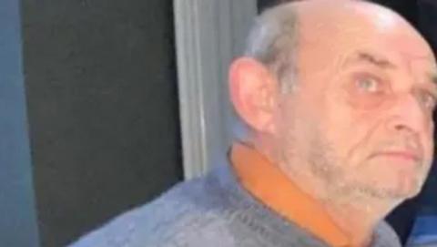 Robert Wilkin is bald and has grey stubble, he is wearing grey jumper and the orange collar of a polo shirt is poking out from beneath