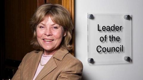 A woman with neck-length brown/blonde hair wearing a pink top and a brown jacket leans against a doorway next to a sign reading "Leader of the Council".