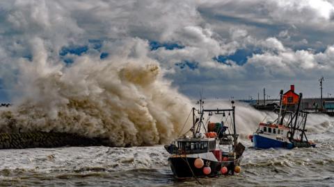 A massive wave crashes off a pier with boast in the water in the foreground and the distance and huge clouds in the sky