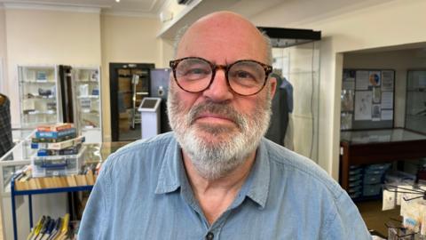 A bald man with thin grey hair on the side of his head. He has a grey beard and moustache and is wearing brown glasses. He is wearing a blue denim-type shirt. Behind him are items on display at the museum.