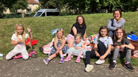 Five young girls sitting on a grassy patch at a playground, with two mums sitting behind them. It is a sunny day and the children are wearing colourful dresses while one is kneeling on a pink scooter. 