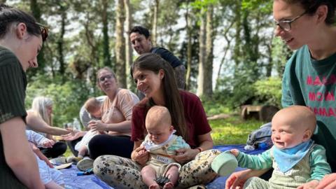 A group of parents and their babies sit in a park on a blue picnic blanket.