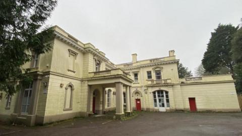 An outside shot of Pounds House located in Central Park in Plymouth. It is a large cream painted building with a canopy covering the front entrance with three red doors. It is surrounded by trees and has plenty of room for parking outside. 