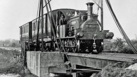 A historic black-and-white photograph showing a steam train crossing a lift bridge on the Hundred of Manhood and Selsey Tramway. The bridge spans a waterway while the locomotive and carriage travel across it.
