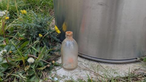 A glass bottle on concrete next to a metallic bin. The glass is opaque and seems to have orange liquid in it. The top of the bottle is crusted with orange paste.