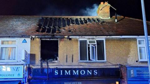 Fire damage to the upper floor and roof of a terraced property. Several roof tiles are missing, exposing charred roof beams, with a window completely removed. The word SIMMONS is printed on hoarding.