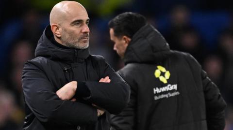 Enzo Maresca, wearing a black hooded coat and looking serious, stands with his arms folded on the touchline
