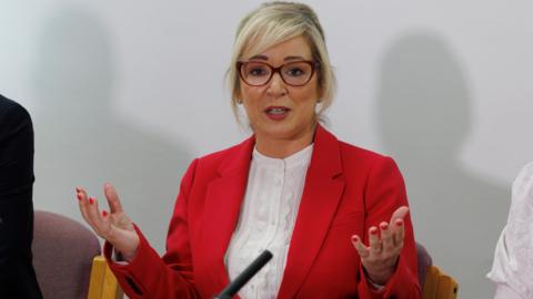 Sinn Fein vice president Michelle O'Neill, during the launch of the party's reform proposals at Parliament Buildings, Stormont. She has blonde hair worn up with red glasses and white shirt and a red jacket. She is mid speech in this picture and holds her hands out in front of her