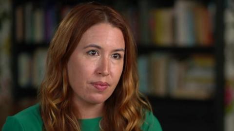 Emily Hunt has long auburn hair and is wearing a green top. She is speaking to a journalist who is off camera as she sits in a sitting room that is out of focus in the background.