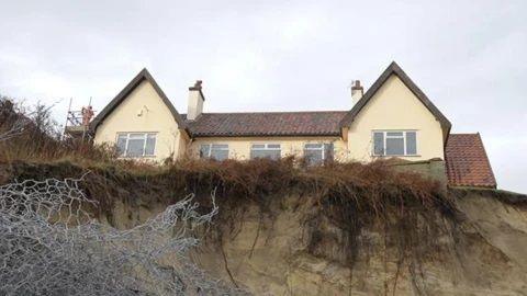 A view of The Chantry property taken from the beach below. Is is a large detached home with two gables at either end. 
There is a sandy cliff that has eroded and is now very close to the house. Rough grass grows over the edge of the cliff, but it is mostly sand. There is wire netting in the foreground.