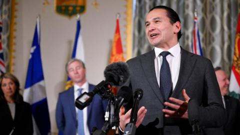 Wab Kinew, Premier of Manitoba, speaks during a press conference with the premiers of Canada in Washington, DC.  He has a slight smile on his face as he speaks into a number of microphones. Behind him stand other Canadian premiers. 