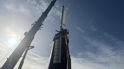 A crane is mid-way through attaching white sails back to a windmill. The windmill has a white cap and black side and the sun is shining on it.