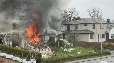House in flames on a street - billowing grey smoke rises from the single-storey white building