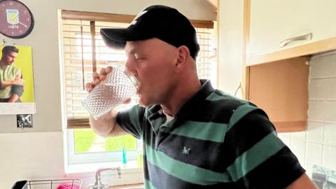 Matt Winter drinking a glass of tap water. He is wearing a striped T-shirt and a cap and is in a kitchen