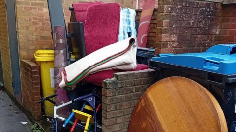 A mattress, table, bicycle, chair and rolled up carpet stuffed into a small communal bin area with brick walls surrounding it.