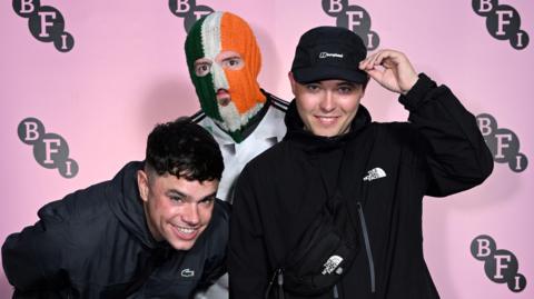 Three young men stand together. One is wearing a green, white and orange balaclava. A pink backdrop is behind them.