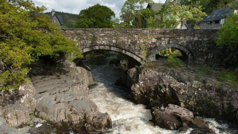 Swallows Falls along the River Lligwy, Snowdonia National Park, Betws-Y-Coed, Wales. It shows white water beween rocks flowing under a stone bridge. Trees and houses are visible in the background