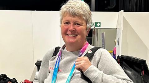 Laura Scarlett-McAllister has short grey hair and is wearing a grey Team England jumper and a large black backpack.