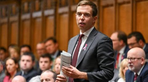 A man in a grey suit with red tie stands in the House of Commons to ask a question. He is holding some folded papers. 