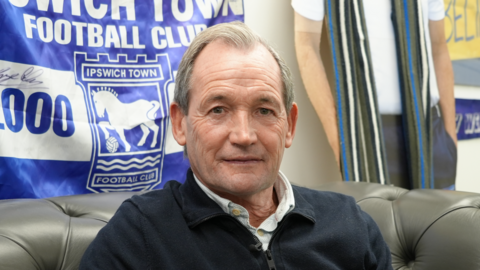 A head and shoulders image of George Burley. He is wearing a navy jumper over a smart shirt. He is sitting on a black leather sofa while smiling and looking into the camera. An Ipswich Town Football Club flag hangs on a wall behind him