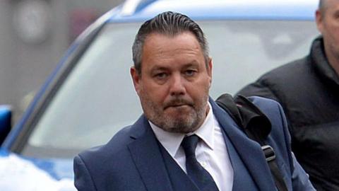 A man with dark hair swept back and a short beard is wearing a blue suit, white shirt and blue tie
