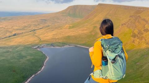 A hiker wearing a backpack looks across at picturesque mountains and a lake in Bannau Brycheinog, also known as the Brecon Beacons