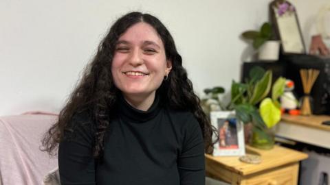 Chiara, a young woman with long, dark curly hair, is sitting down and smiling. She is wearing a black turtleneck top. In the background is a sofa and a table with a plant and a picture on.