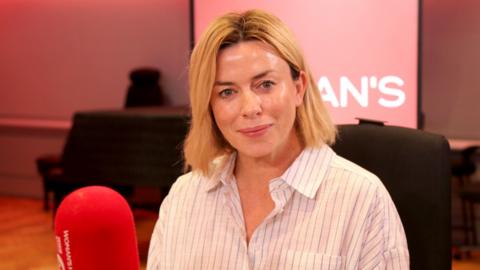 Eve Myles sits in a radio studio behind a red mic. She has shoulder length blonde hair and wears a white shirt
