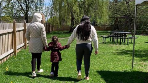 Aracely and her two daughters link hands in a park - we only see them from their back