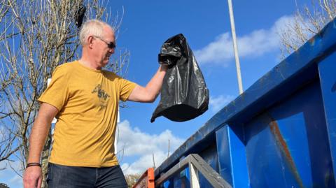 Man with sunglasses wearing a mustard yellow top and jeans is putting his black bin back of rubbish into a skip