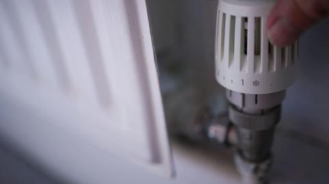 A hand turning a white thermostat on a radiator down to its lowest setting.