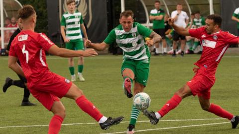 A group of footballers go for the ball. Two of them are wearing a red strip but the player in the centre who has got the ball is wearing a green and white strip.