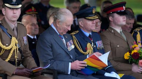 The king sits in the front row of the dedication ceremony flanked by uniformed members of the armed forces. He is looking down reading an open book of service with the LGBT rainbow on the front.