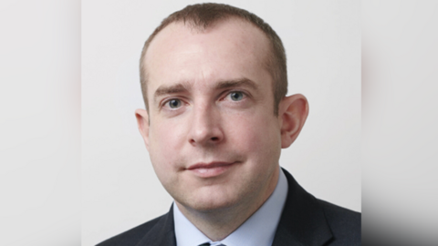 James Blythe is sat in front of a grey background. He is wearing a dark jacket, blue shirt and tie, which is hidden from the picture. He has short brown hair.