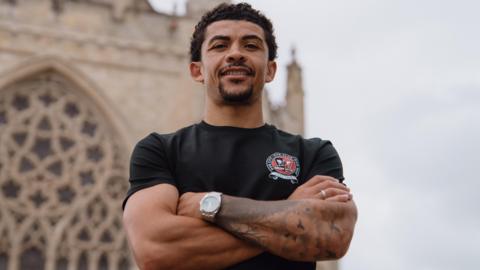 Josh Gordon, wearing a black tee-shirt and folding his arms, is smiling while standing outside Exeter Cathedral