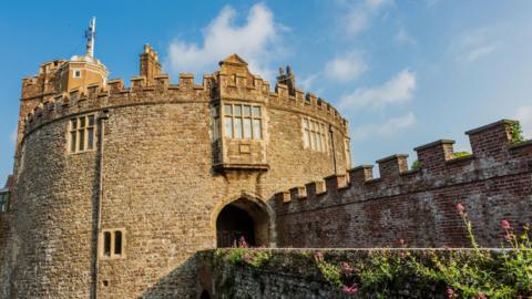 A sunny day with blue skies and a round stone castle in the Tudor style with pink flowers growing out of a brick bridge leading to the castle archway