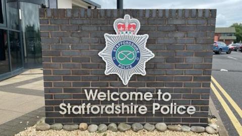 A brick wall outside Staffordshire Police headquarters. The wall has the force's badge on it, a silver star shamed symbol with a red crown and a silver rope on a green background, and the wording 'Welcome to Staffordshire Police'
