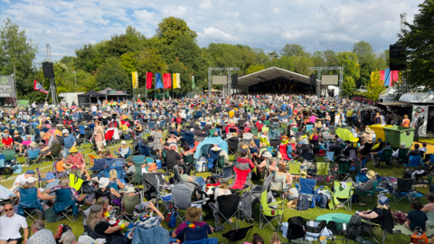 A crowd of people, many sitting on camping chairs, in front of an outdoor stage in a park 