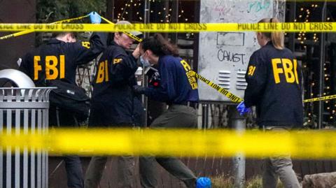 Yellow police tape is in the foreground partially covering four FBI agents combing through a crime scene.