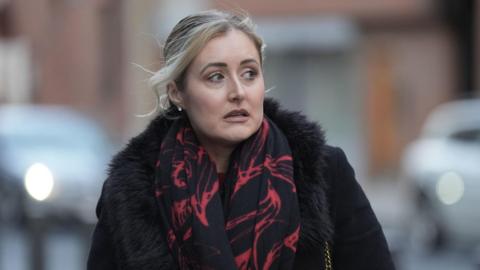 A woman wearing a black coat and a black and red patterned scarf walks through a London street.