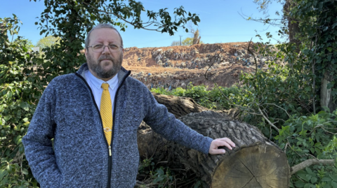 Councillor Carubia stands in front of the rubbish tip from a nearby field. He is wearing a shirt and yellow tie under a fleece. His arm rests on a tree stump.