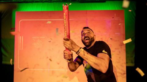 A man wearing a black T shirt holding a confetti canon and smiling while he looks into the sky