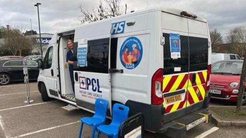 A woman in blue scrubs stands in a white medical vehicle with NHS logos and branding on it. The woman has blonde hair and smiles at the camera. The vehicle is parked in a car park with the door open and two blue plastic chairs in front of it.