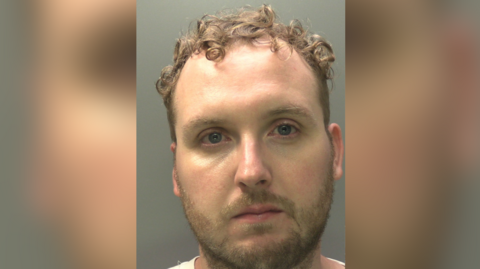 Craig Winston is wearing a white t-shirt. He has short cropped facial hair and curled brown hair.