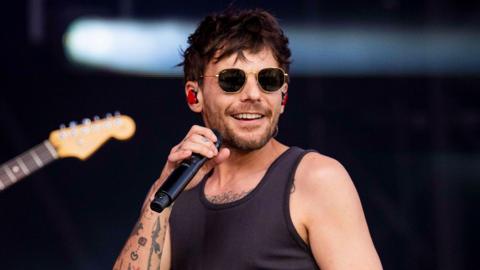 Louis Tomlinson, wearing a black vest and sunglasses, holding a microphone and performing at Radio 1's Big Weekend