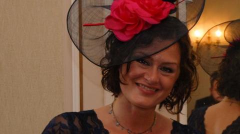 Maria Morris, who died at Bethlem Royal Hospital in 2021, is standing in front of a mirror at a wedding smiling. She is wearing a black dress, and a black and pink fascinator. 