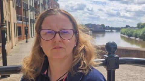 A head and shoulder shot of Su Roys, who has has shoulder-length brown hair, wearing a navy England Rugby polo shirt and burgundy-rimmed glasses. She is stood on a path in front of the River Trent. The bridge can be seen in the distance.