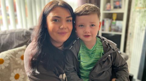 Catherine is sitting in her living room with Séadhna on her lap. They are both wearing grey. They are sitting with their backs to the window. There is a daisy decoration on a cushion. 