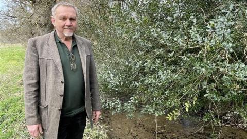 Alex Gater standing next to a leat which has been contaminated by raw sewage. He is looking at the camera and is wearing a grey suit jacket with a green shirt. The leat has stones at the bottom and there is a large branch, with green leaves, hanging over the top of it.